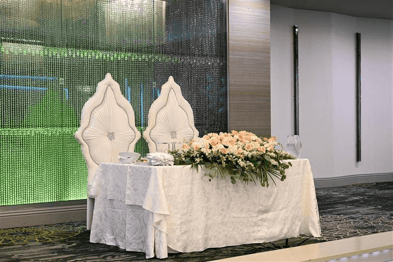 Elegant wedding setup with white chairs, floral arrangements, and a decorated table against a shimmering backdrop.