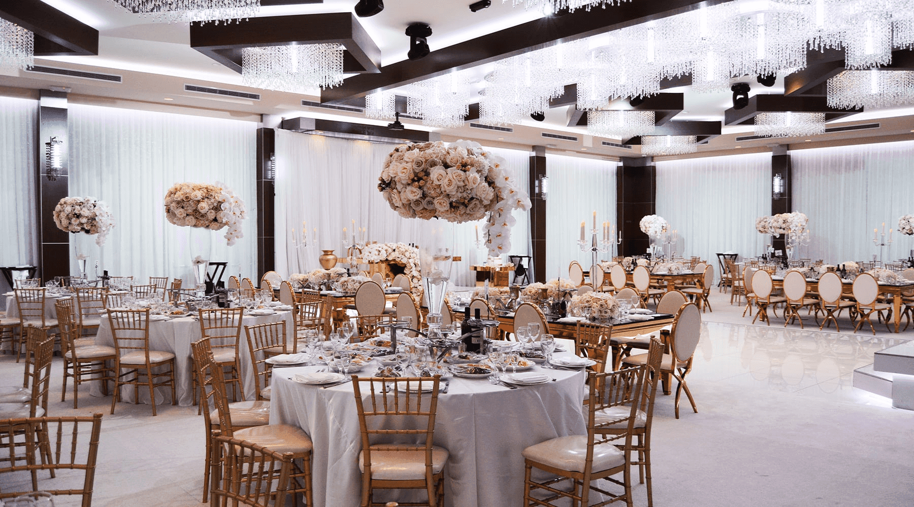 Elegant banquet hall with white and gold decor, oversized flower centerpieces, and sparkling chandeliers. Perfect for events.