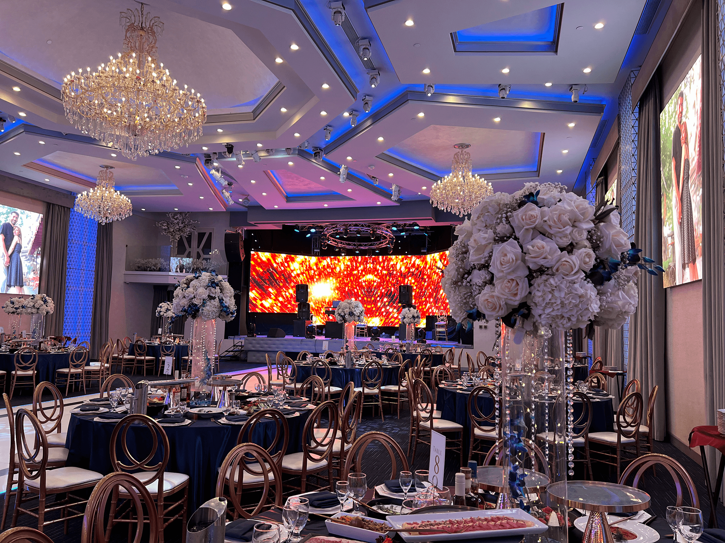 Elegant banquet hall with chandeliers, floral centerpieces, and a vibrant stage backdrop, set for an event.