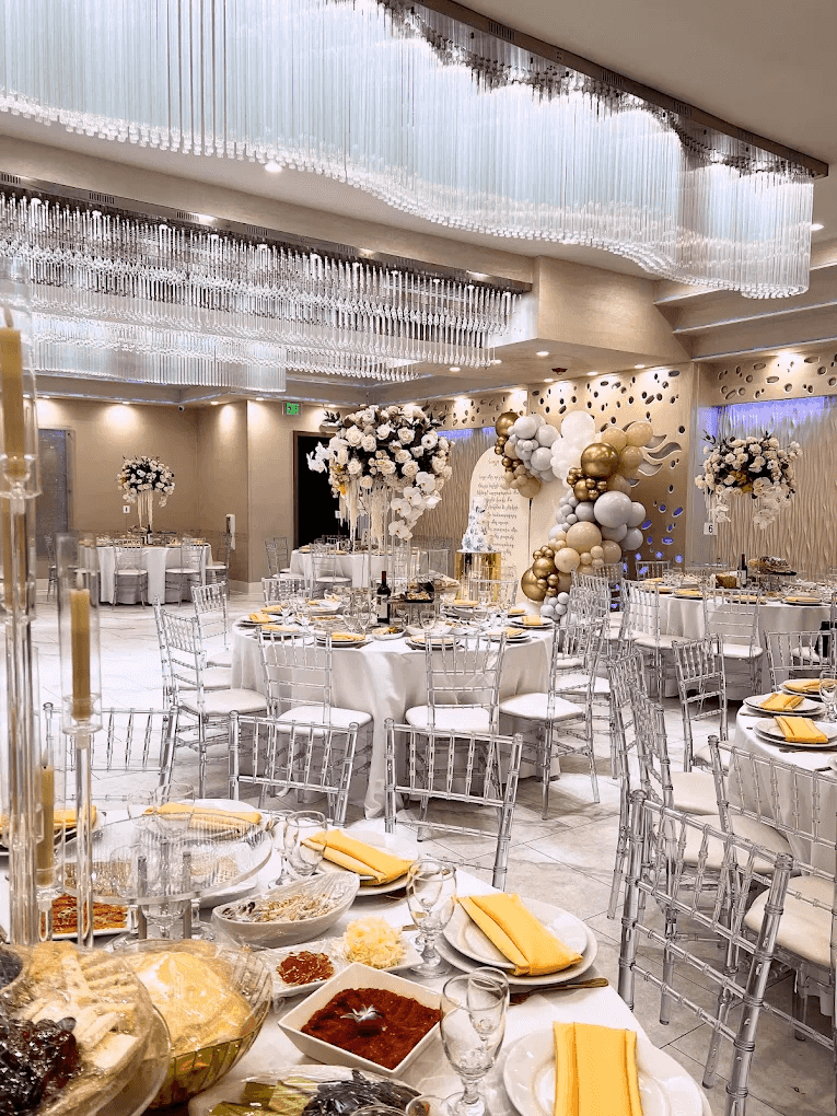 Elegant banquet hall with silver chairs, decorated tables, floral arrangements, and a lavish buffet of dishes and desserts.