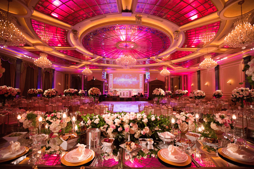 Elegant banquet hall adorned with pink and gold decor, featuring floral centerpieces and crystal chandeliers.