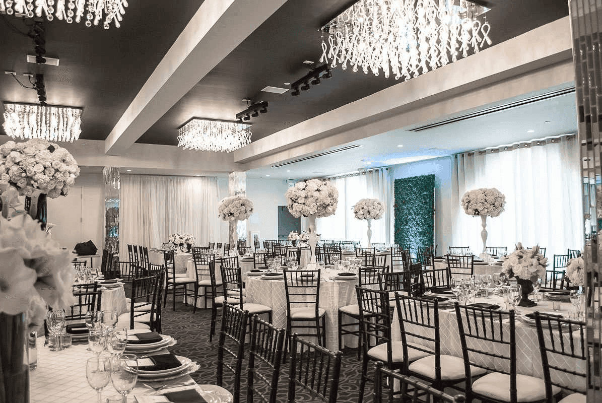 Elegant banquet hall with crystal chandeliers, white floral centerpieces, and black chiavari chairs, set for a formal event.