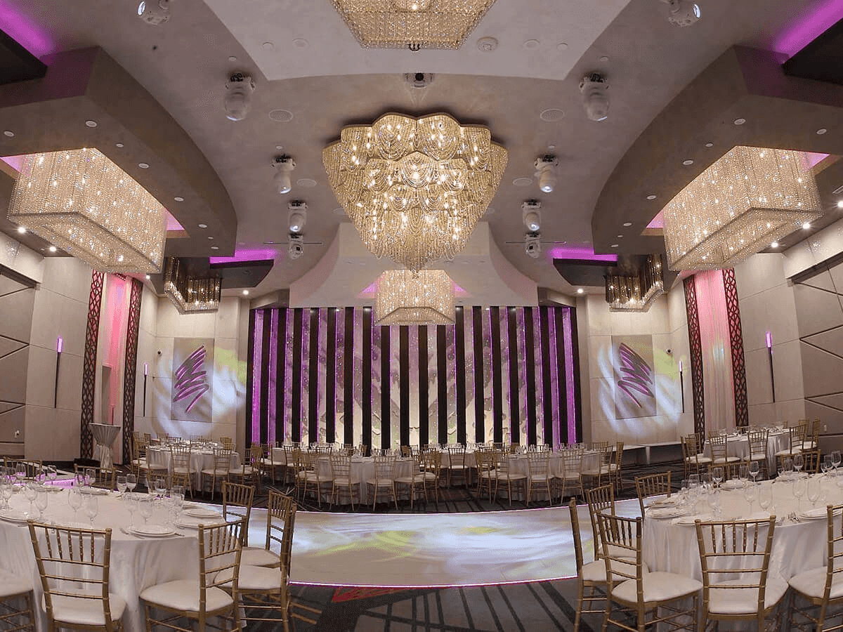 Elegant banquet hall with gold chandeliers, white tables, pink lighting, and a floral backdrop, ready for a celebration.