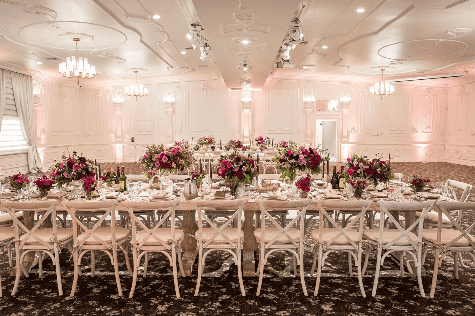 Elegant banquet setup with pink floral centerpieces, white wooden chairs, and soft lighting in an ornate room.