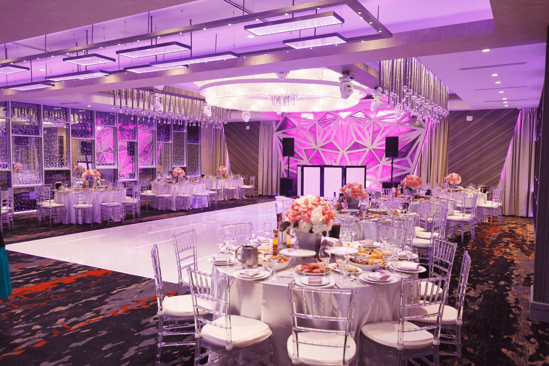 Elegant banquet hall with pink lighting, round tables, silver chairs, floral centerpieces, and a dance floor.
