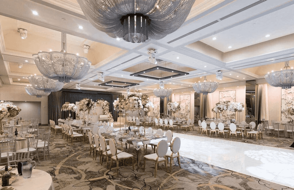 Elegant banquet hall with golden accents, white chairs, floral decor, and stunning chandeliers, perfect for celebrations.