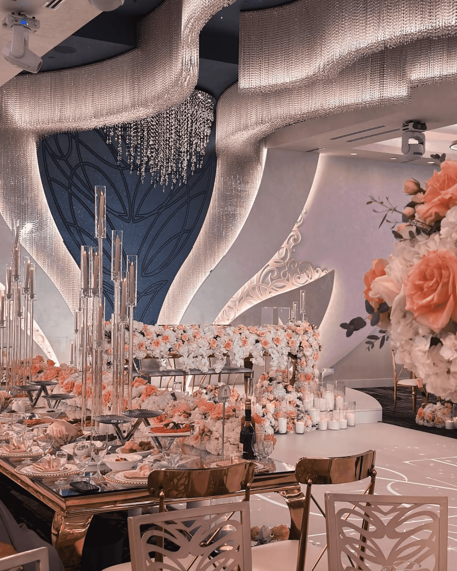 Elegant banquet hall with white and peach floral decor, crystal chandeliers, and lavish table settings for an event.