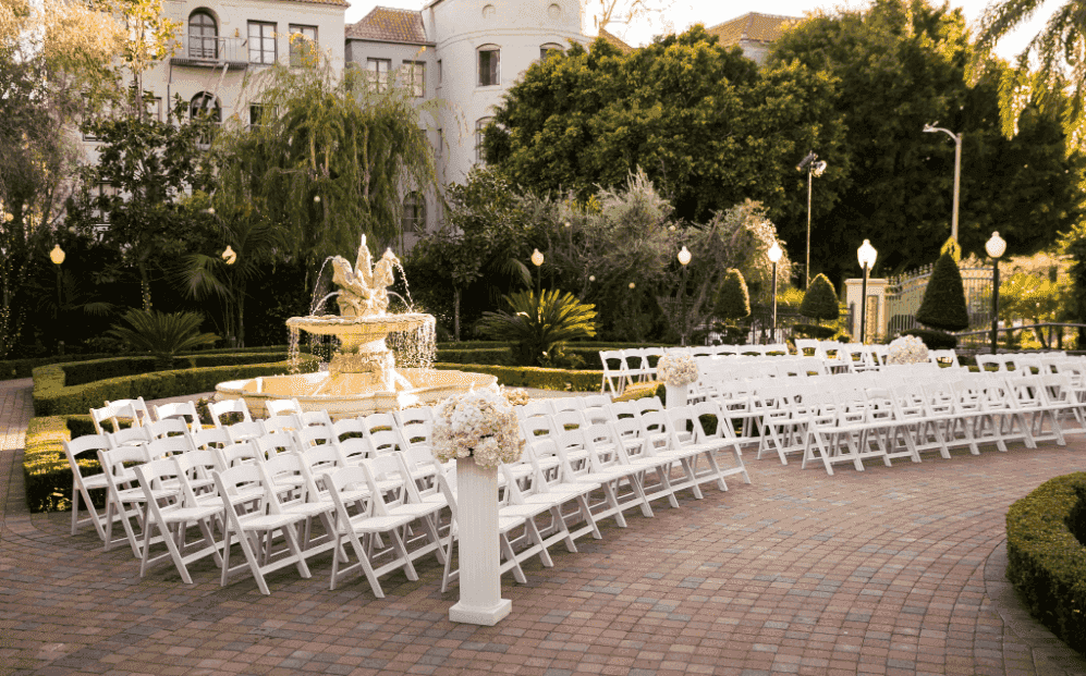 Elegant outdoor wedding venue with white chairs arranged around a classic fountain, surrounded by lush greenery.